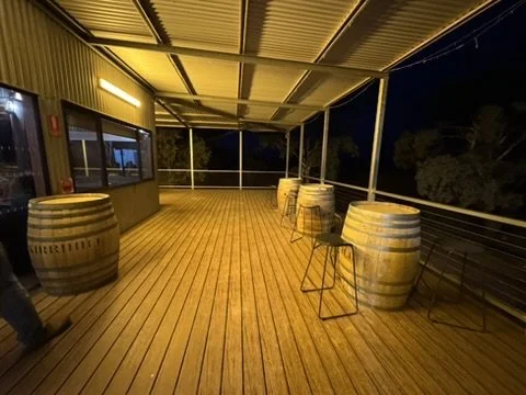 Empty outdoor patio with wooden flooring, large barrel tables, and a few metal chairs under a yellow roof at night.