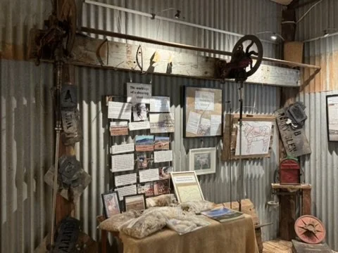 A rustic interior with corrugated metal walls, displaying vintage tools, photographs, and posters, along with a wooden table covered with fabric and books, and various antique items and décor.