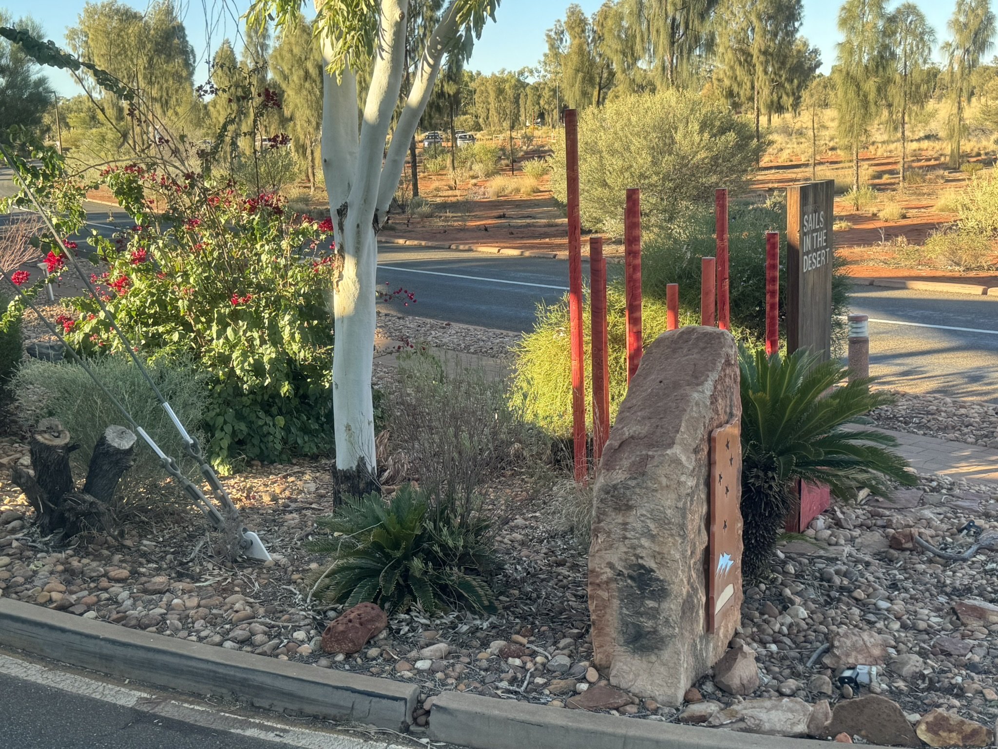 Desert landscaping with a tree, bushes, rocks, and red-painted posts along a roadside. There is a sign that reads 'Sails in the Desert' and a large rock with decorative art on it.
