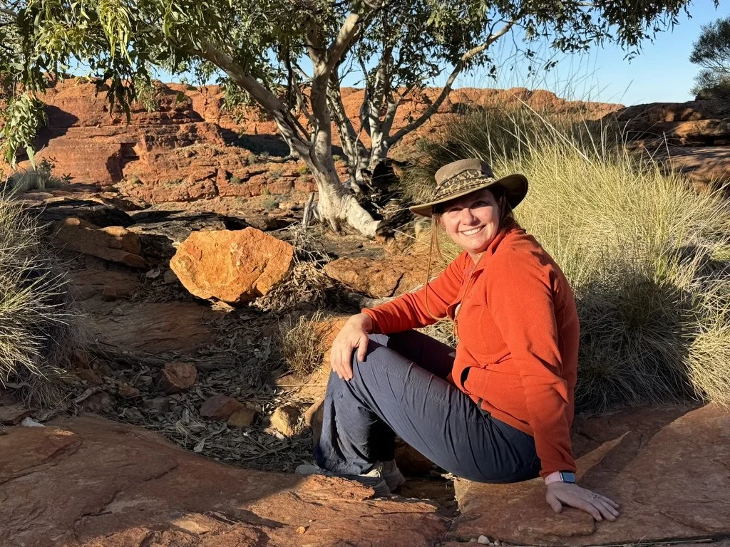 A woman sitting on a large rock in a desert landscape, wearing an orange jacket, gray pants, and a wide-brimmed hat, smiling at the camera with desert vegetation and red rock formations in the background.