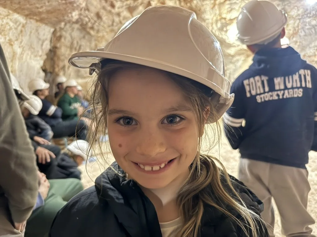 A young girl with a big smile, wearing a white hard hat, at an underground cave tour with other visitors also wearing hard hats in the background.