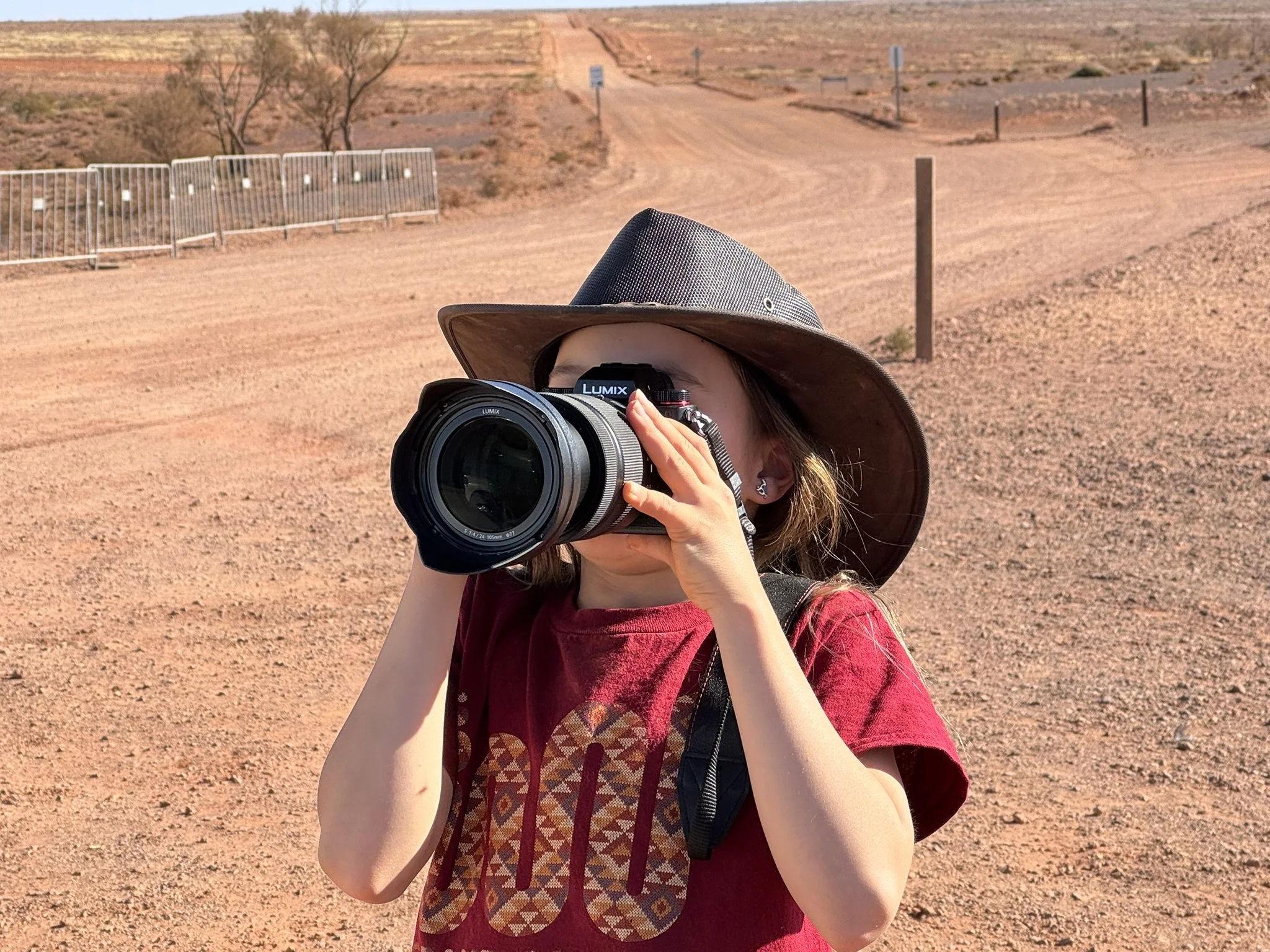 Young girl wearing a wide-brimmed hat taking a photograph with a camera in a desert landscape.