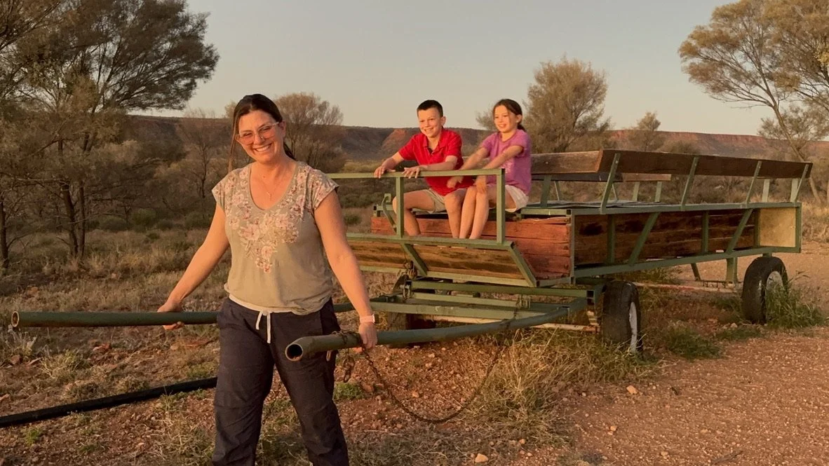 A woman pulling a wooden farm wagon with two children sitting inside, in a rural outdoor setting during sunset.