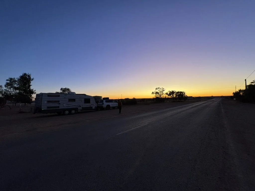 Sunset over a rural dirt road with a travel trailer and a white vehicle parked nearby, trees in the background.