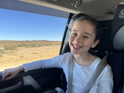 A young girl with a big smile sitting in a vehicle seat, looking out the window at a clear, open landscape with blue sky and dry land.