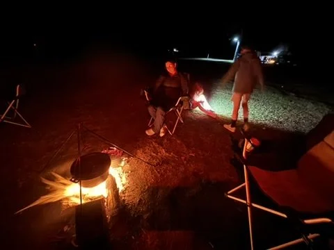 People sitting and standing around a campfire at night, with outdoor chairs and a grassy area nearby.