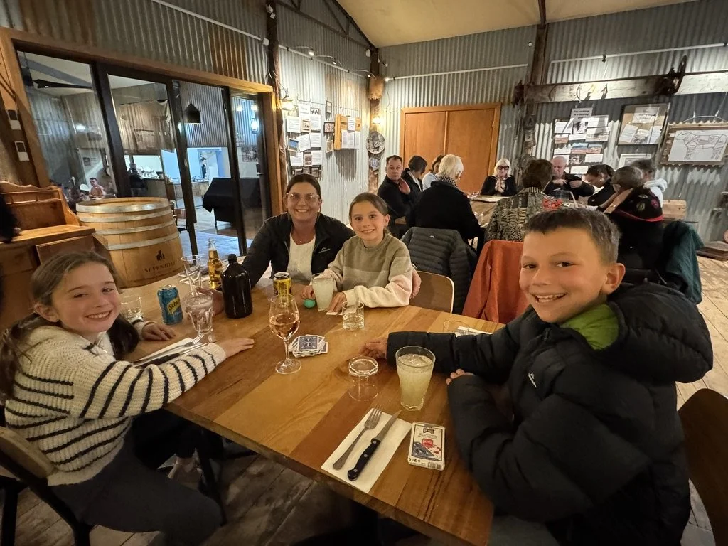 Four children and a woman sit at a table in a rustic restaurant, smiling at the camera. Other diners are visible in the background.