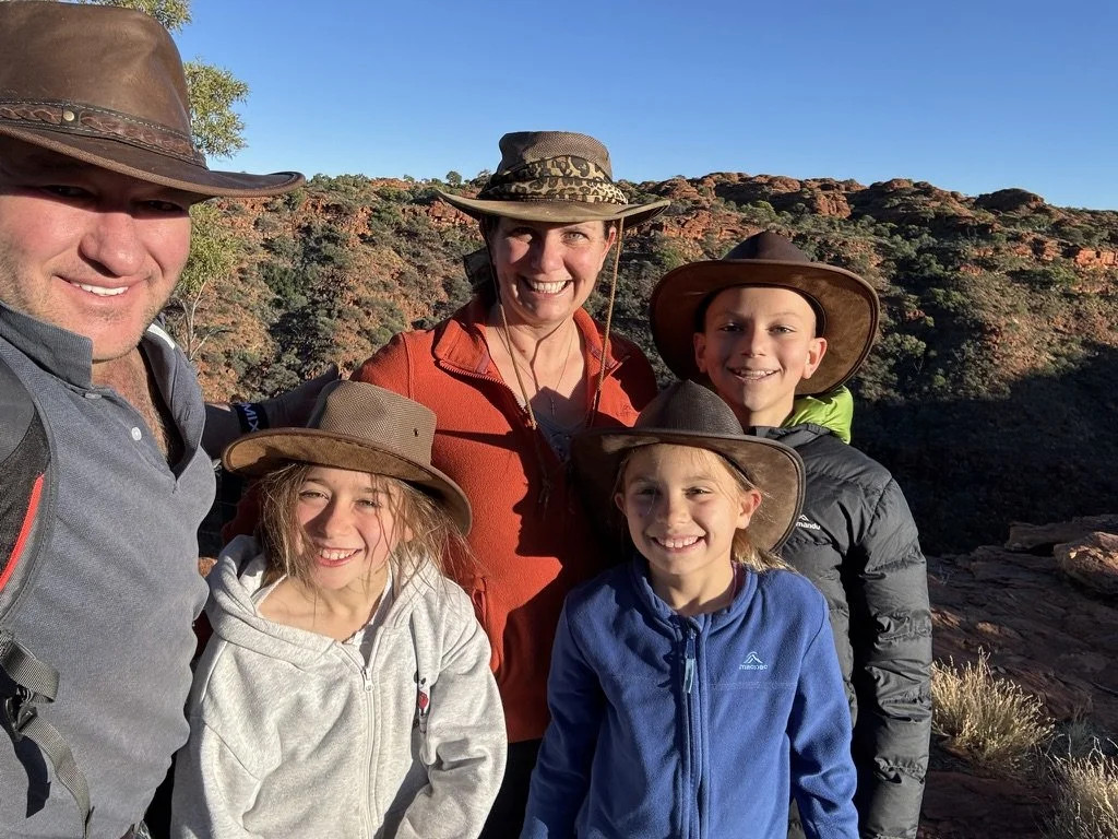 A group of six people, including two adults and four children, dressed in outdoor hiking gear, smiling for a photo during a hike in a rocky, desert terrain with sparse vegetation and clear blue sky in the background.