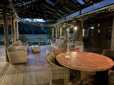 Covered outdoor patio with hanging string lights, wicker chairs, a round wooden table, and a view of a field.