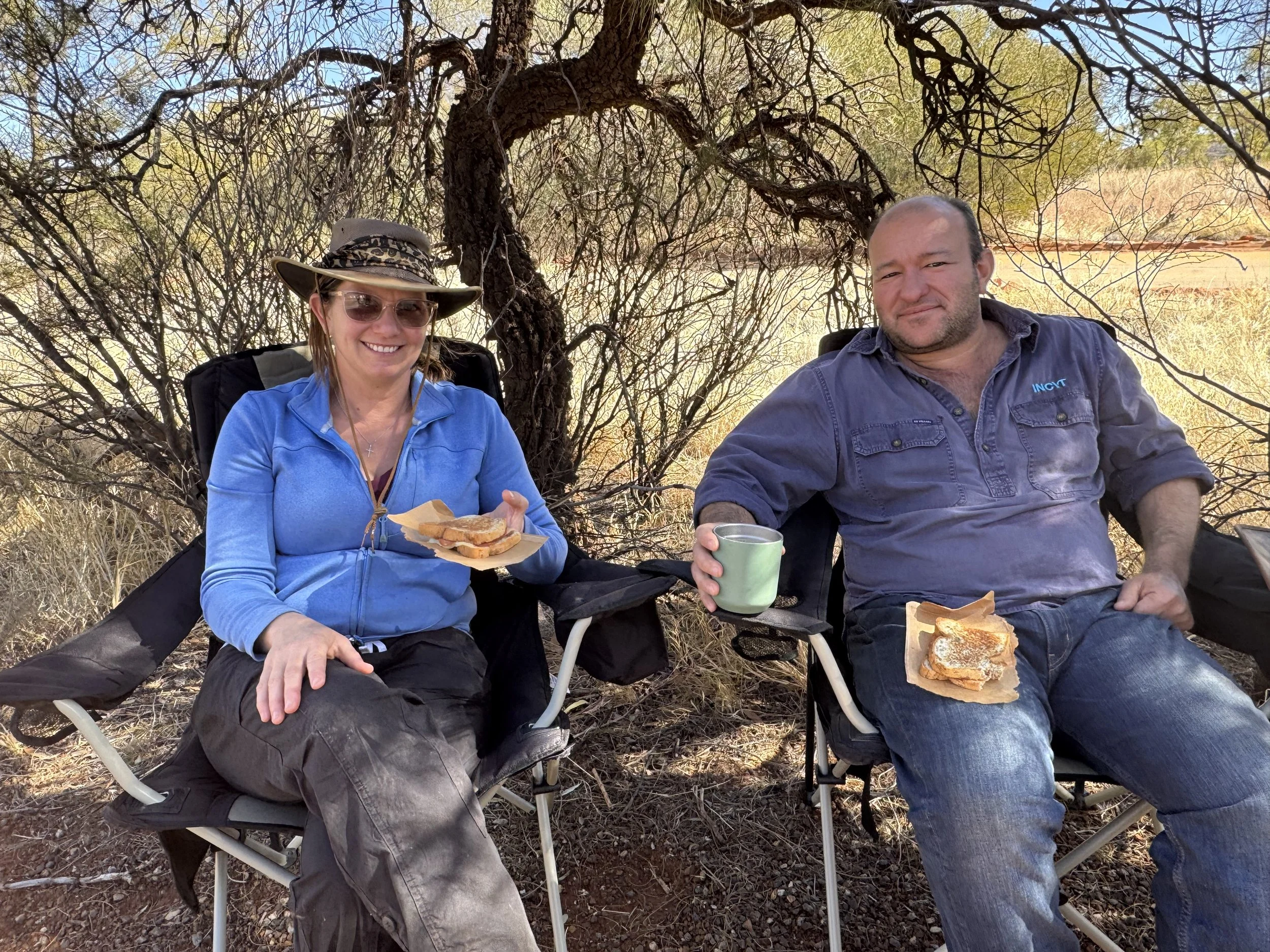 A woman and a man sitting outdoors in camping chairs, enjoying sandwiches and drinks, with a tree and desert landscape in the background.