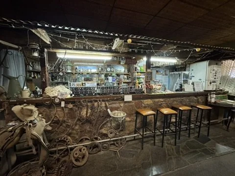 Interior view of a rustic bar or café with a wooden counter, four bar stools, and vintage decor including an old bicycle, hats, and various knick-knacks.