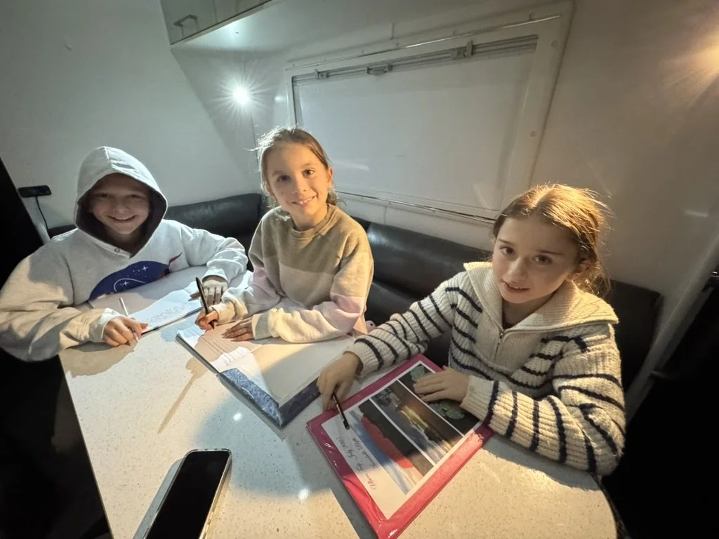 Three children sitting at a white table, smiling and engaging with notebooks and a large photo album, in a cozy indoor setting with a black cushioned bench in the background.