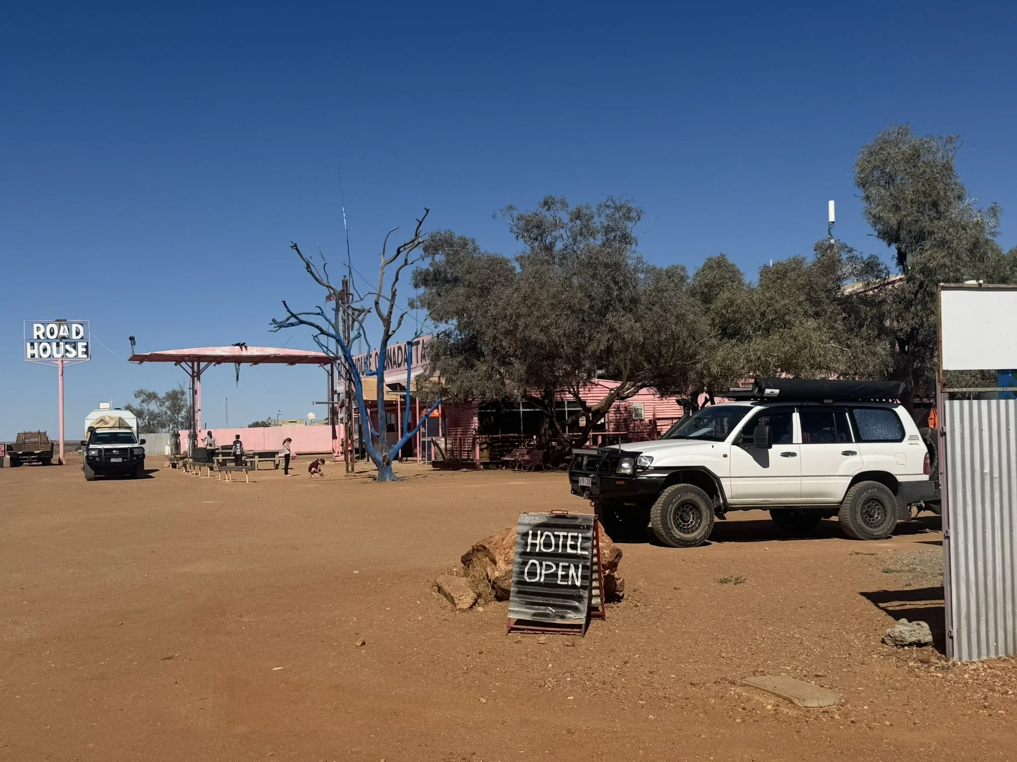 A dusty outdoor area with a white SUV parked beside a sign that says 'Hotel Open'. In the background, there are trees, a pink building, and a wooden structure with benches. A few people are visible near the benches and structures, and a sign on the l