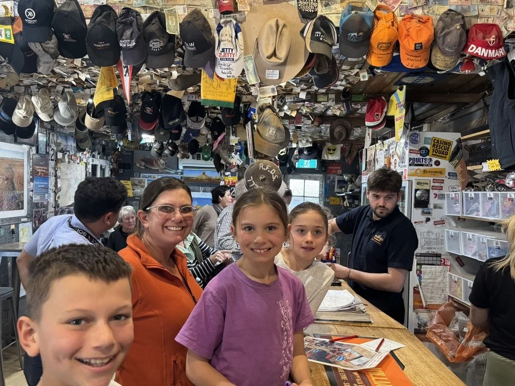 Children and adults inside a store with various hats hanging from the ceiling and signs on the walls, including a counter with papers and a person assisting customers.