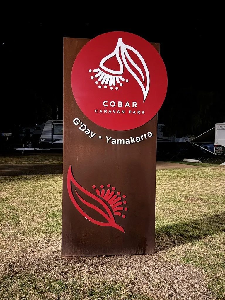Sign for Cobar Caravan Park featuring a red circle with a white floral logo, and a stylized red leaf and floral design on a dark background at night.