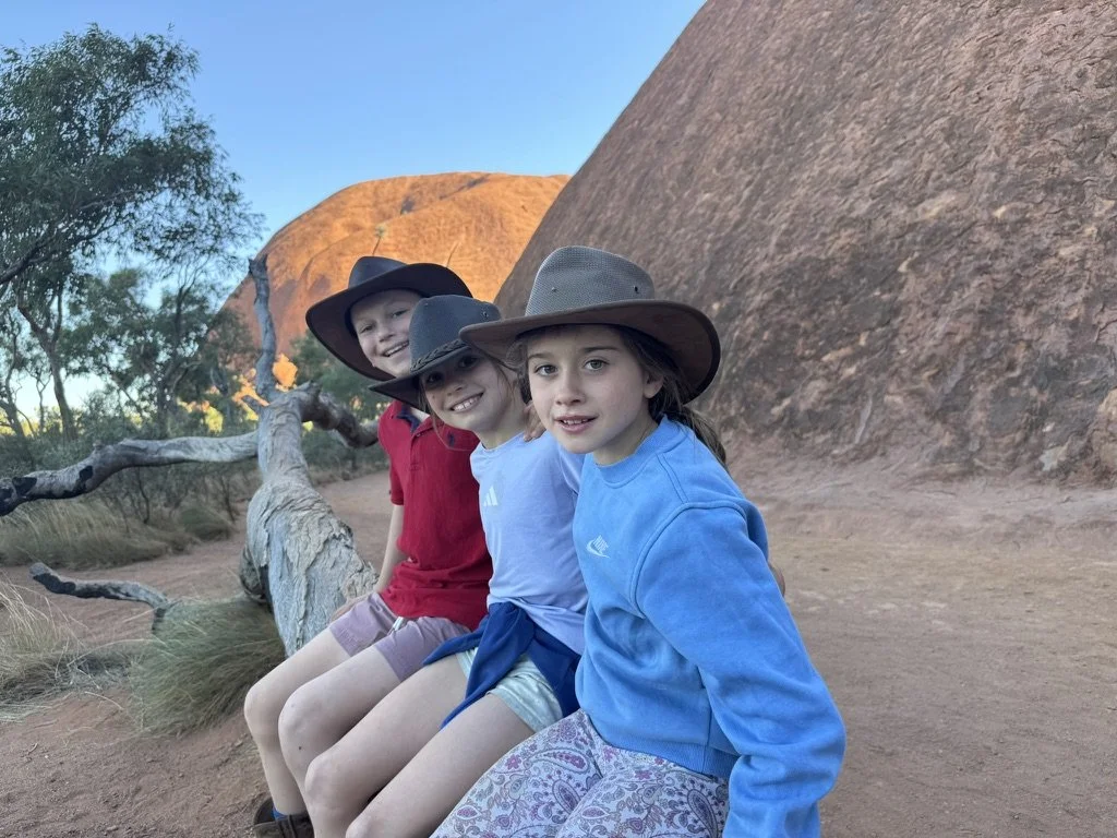Three children sitting on a fallen tree trunk outdoors in a desert landscape, with large rocky formations and sparse vegetation in the background. All three wear wide-brimmed hats and casual outdoor clothing.