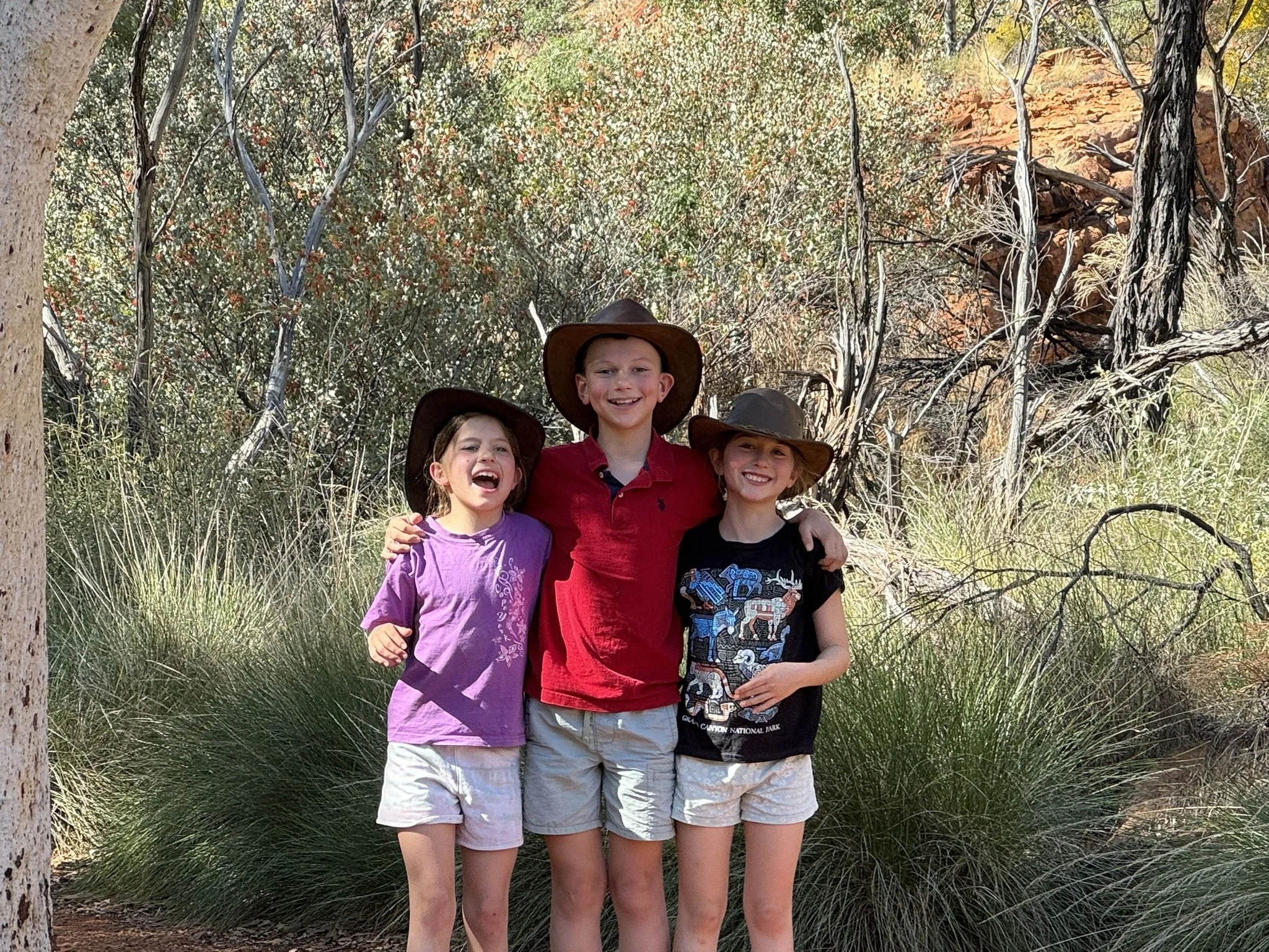 Three children standing close together outdoors in a wooded area, smiling and wearing cowboy hats, with greenery and trees in the background.