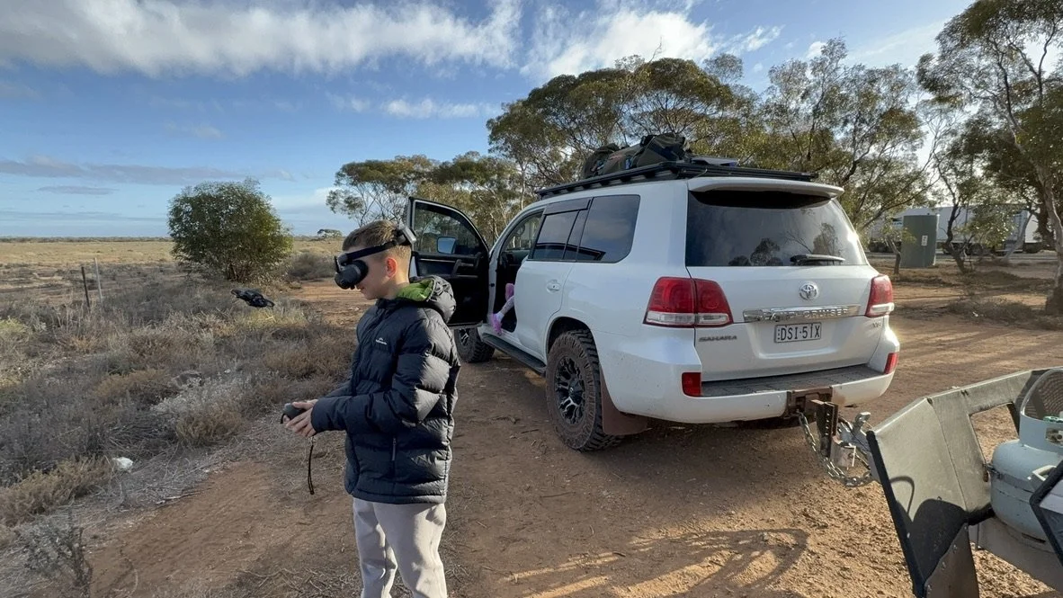 A boy in a black jacket with headphones standing outdoors on a dirt path, holding a remote control, with a white SUV nearby. The SUV is parked with its door open, and it has a surfboard on top and a trailer attached. The landscape includes sparse tre