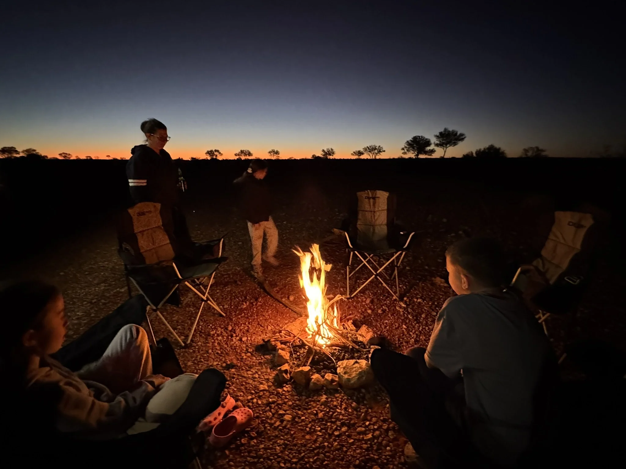 People sitting around a campfire at sunset in a remote outdoor location with silhouetted trees in the background.