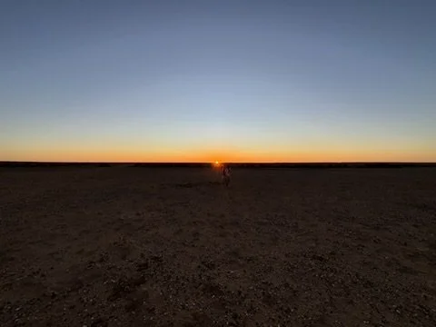 A person walking in a vast, empty desert at sunset with a clear sky.