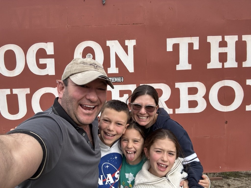 A group of six smiling people taking a selfie in front of a red wall with large white text that reads 'On the...' with part of the text obscured.