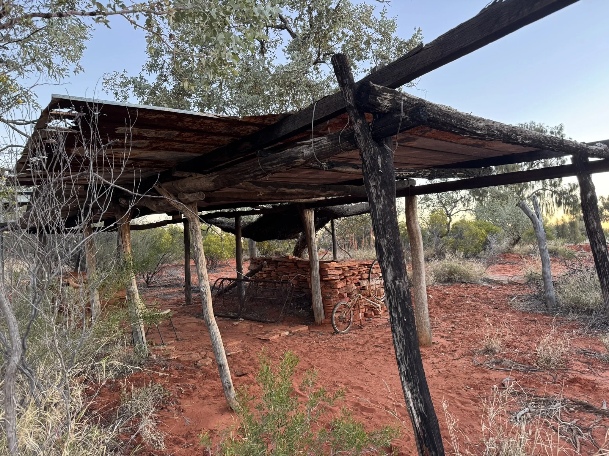 A rustic, weathered wooden shelter with a tin roof in a dry, red desert landscape. There is a brick wall and a rusty bicycle underneath the shelter, surrounded by sparse desert vegetation and trees.