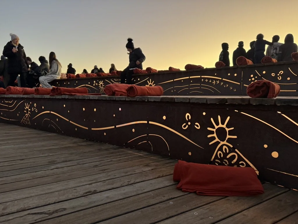 People gathered on a wooden deck at sunset, sitting and standing along a black railing decorated with gold celestial patterns, with red blankets or cloths placed on the railing and on the ground.