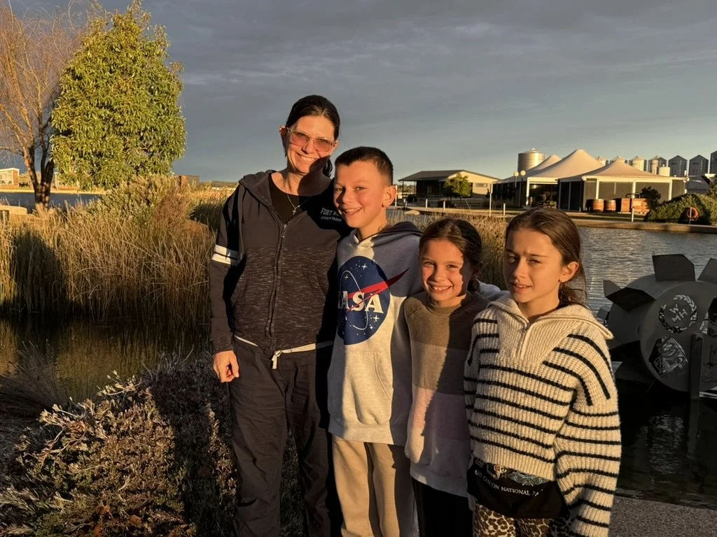 A woman and three children standing by a body of water during sunset with trees, buildings, and tents in the background.