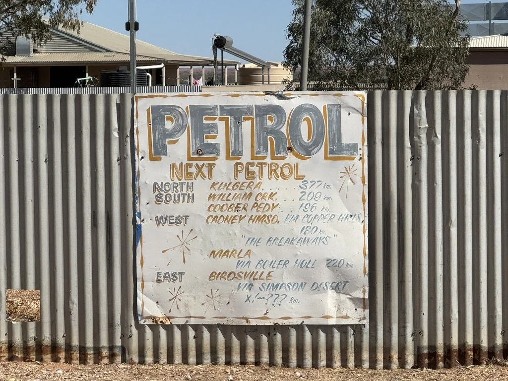 A white sign with handwritten text advertising petrol station locations and distances, attached to a corrugated metal fence.