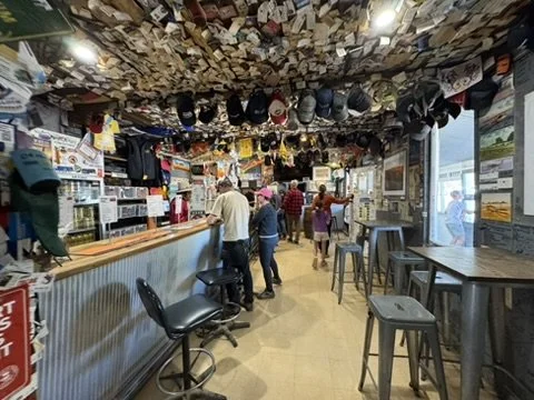 Bar interior with a ceiling covered in dollar bills, people standing and sitting, bar counter with stools, wall decorated with pictures and signs, and large windows letting in natural light.