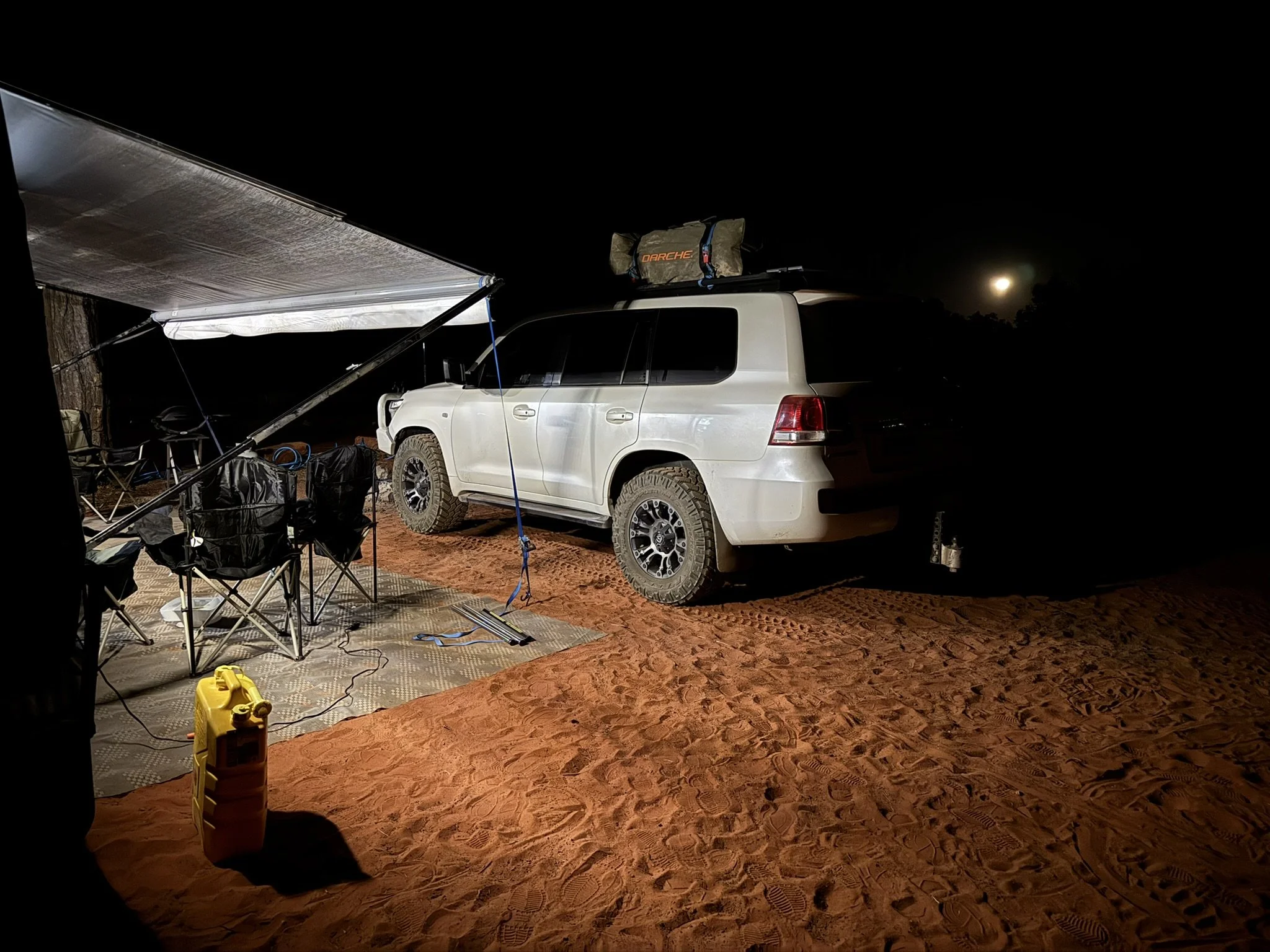 A white SUV parked on red sand at night with a moon in the sky. There is an outdoor setup with folding chairs, a table, and a yellow container on the sand. An awning covers part of the area, and camping gear is on the roof of the vehicle.
