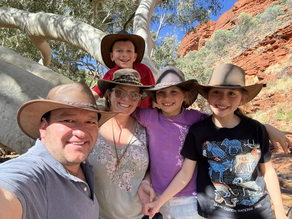A family of five, wearing hats, is taking a selfie outdoors in a desert landscape with red rocks, sparse desert vegetation, and a clear blue sky in the background.