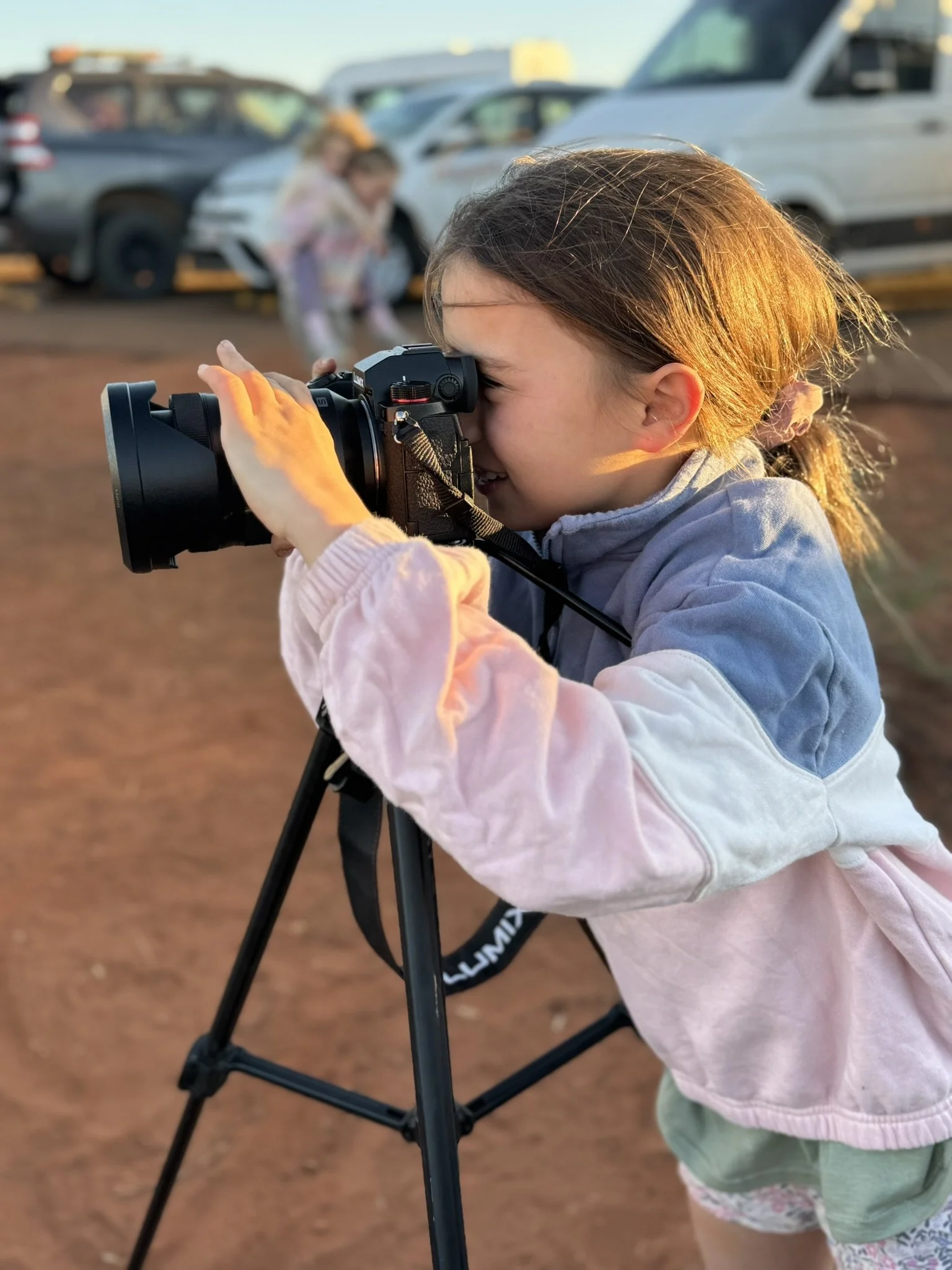 A young girl with brown hair, wearing a pink and blue jacket, is looking through a camera mounted on a tripod, taking a photograph. In the background, there are parked cars and a few children, with the setting illuminated by warm sunlight.