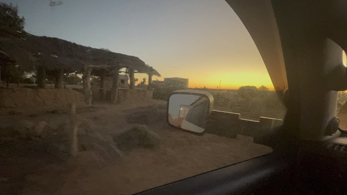 View of a sunset seen through the window of a vehicle, with a side mirror reflecting part of the road and surroundings, and sparse structures and landscape outside.