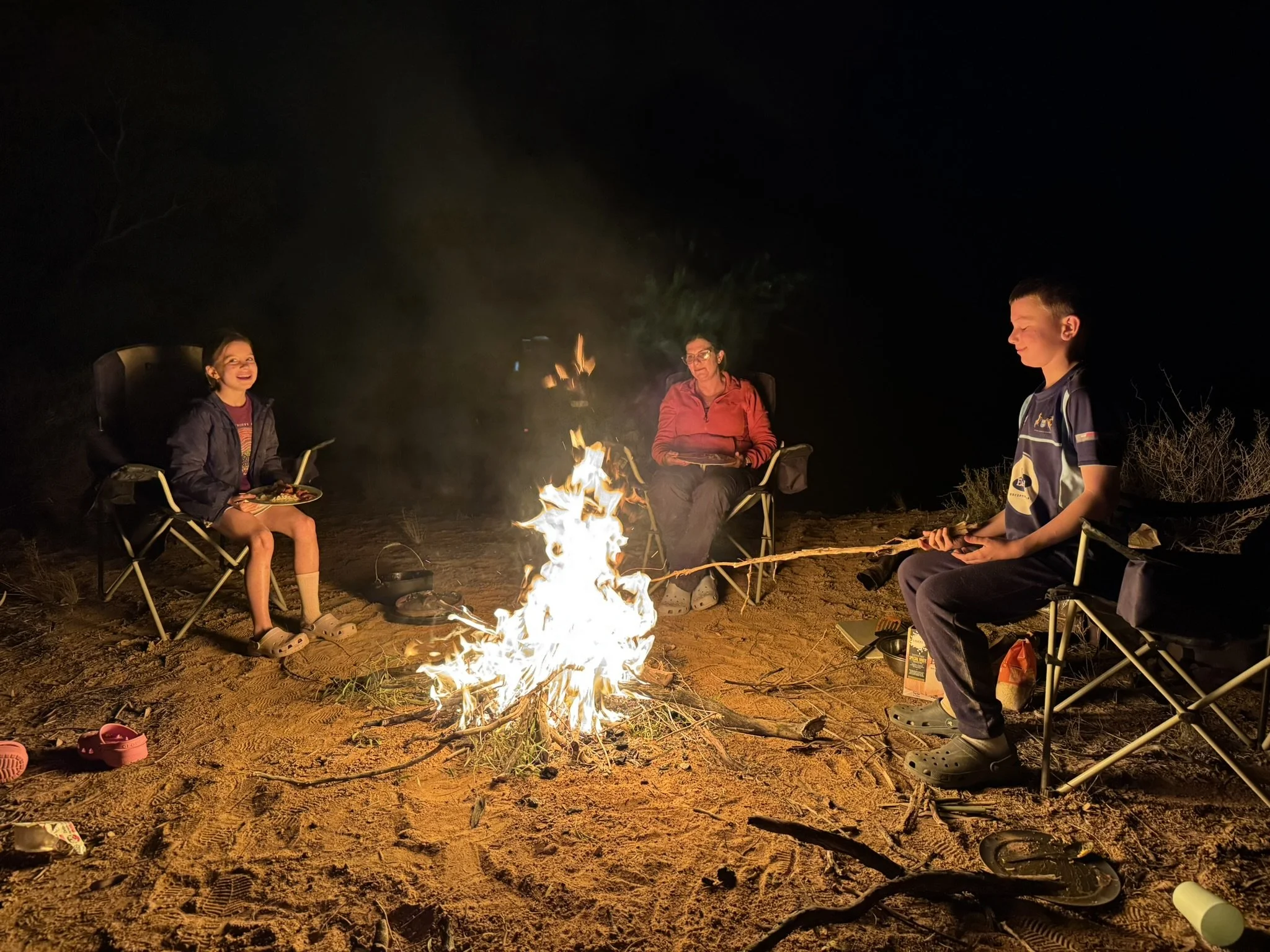 Three people sitting around a campfire at night, in camping chairs, on a sandy ground with some bushes in the background. One person is roasting a marshmallow, while another has food on their plate, and the third is holding a stick with a marshmallow