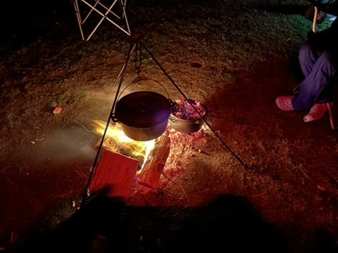 Bonfire with pot hanging over it on tripod and food in bowl, nighttime outdoor scene.