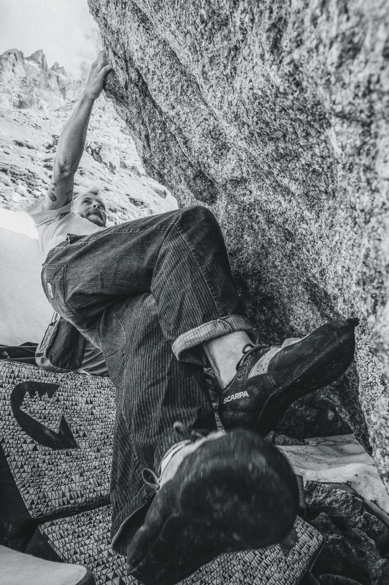 Person climbing a rock wall, gripping a large boulder with both hands, wearing climbing shoes, jeans, and a t-shirt, with other rock formations and climbing gear visible.