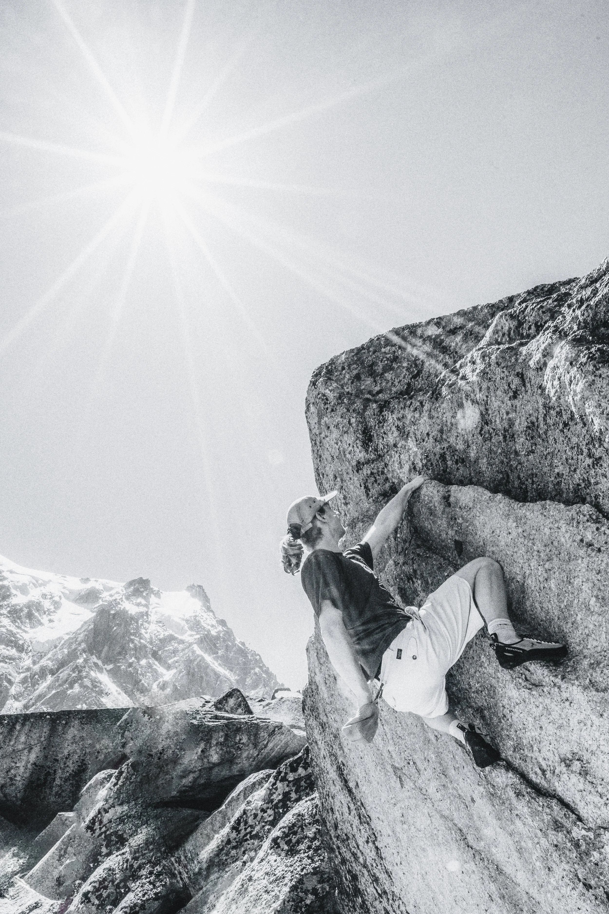 A person rock climbing on a large boulder in a mountainous area during the daytime, with snow-capped peaks in the background and the sun shining brightly in the sky.