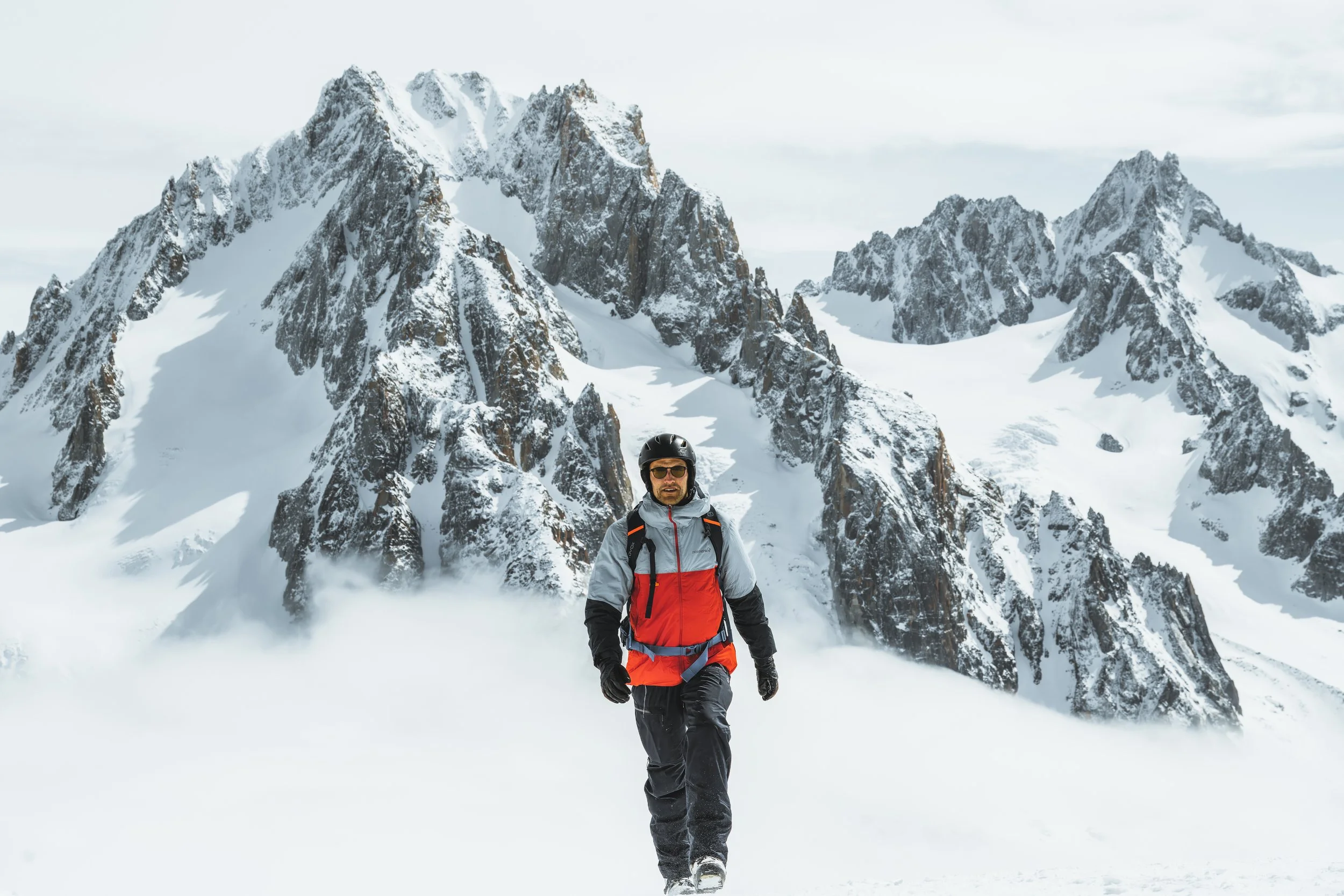 A man wearing a black helmet, sunglasses, and a red and gray winter jacket snowshoeing in a snowy landscape with towering snow-covered mountains in the background.