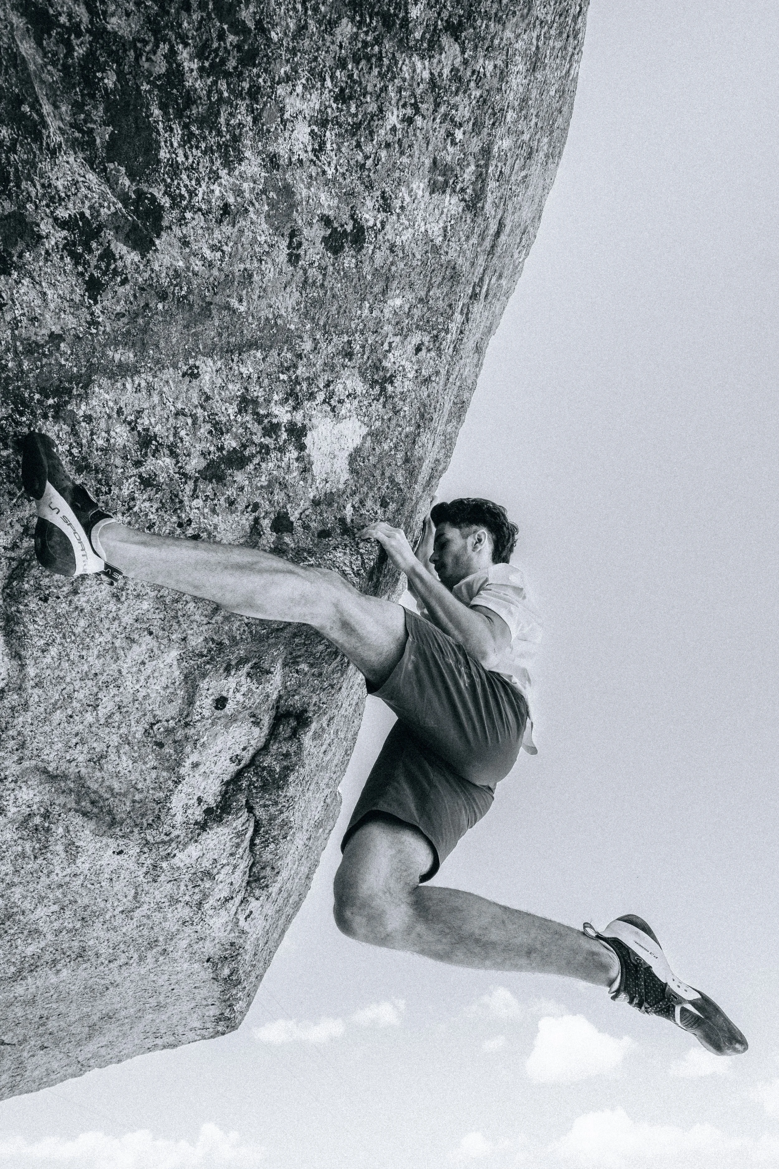 A man rock climbing a large boulder outdoors, wearing shorts, a short-sleeved shirt, climbing shoes, climbing harness, and gloves.