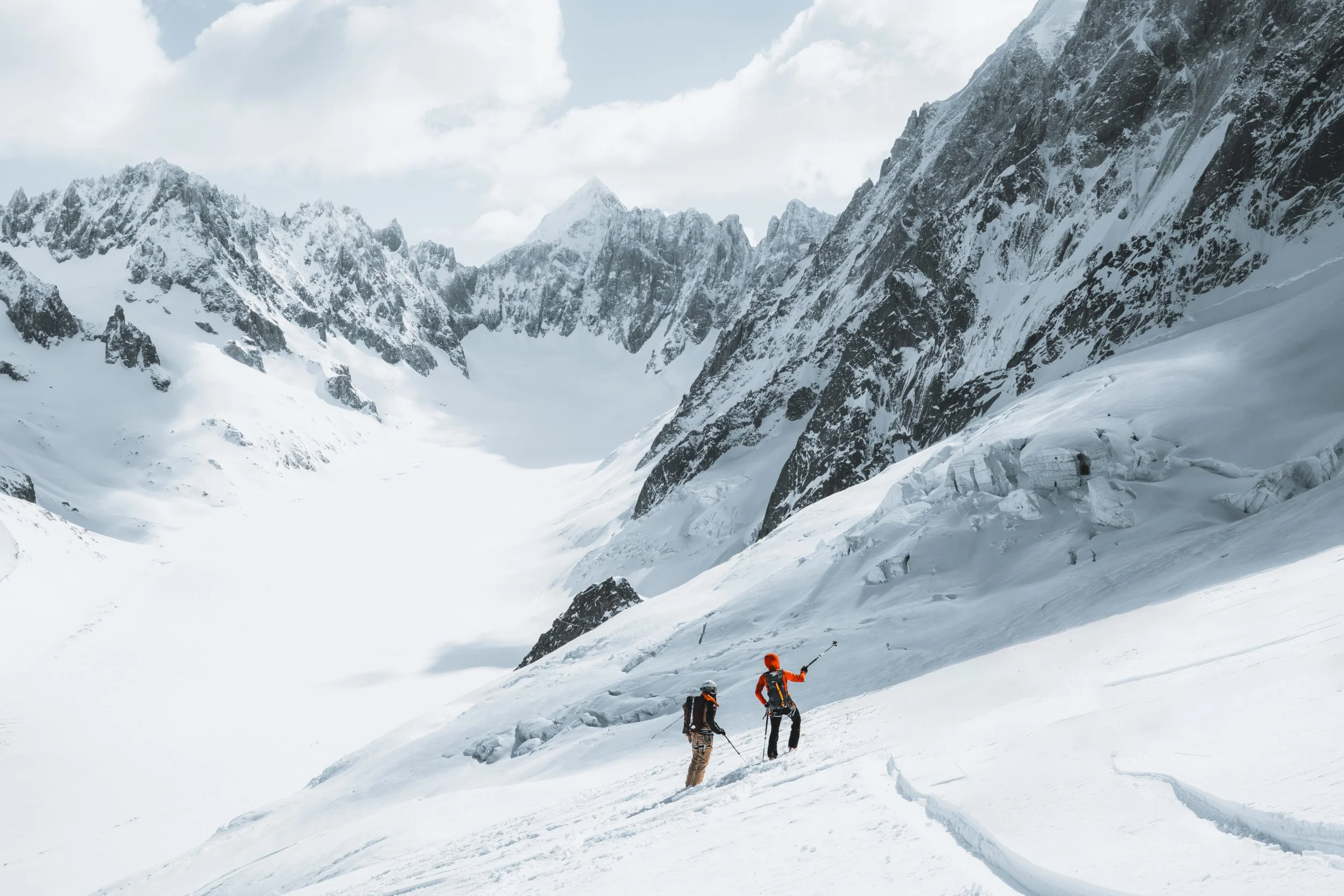 Two climbers with orange helmets and gear ascending a snow-covered mountain in a remote, icy landscape with rugged peaks.