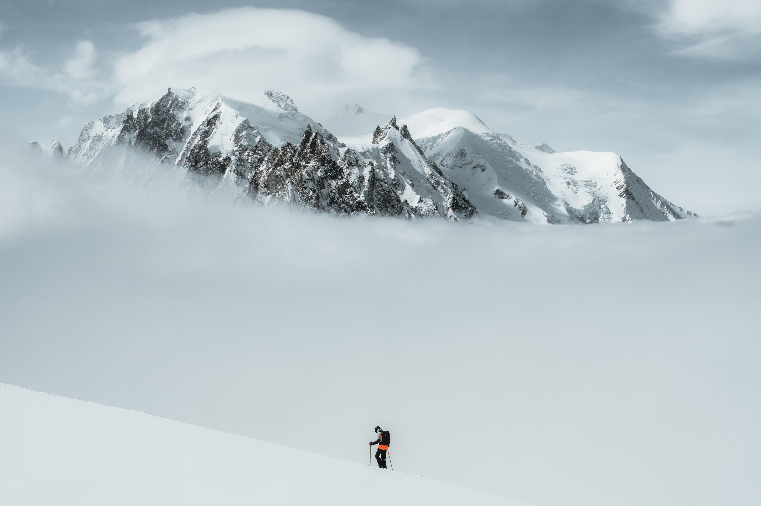 A person wearing winter gear, including a backpack, hiking through snow in a mountainous area with snow-covered peaks in the background under a cloudy sky.