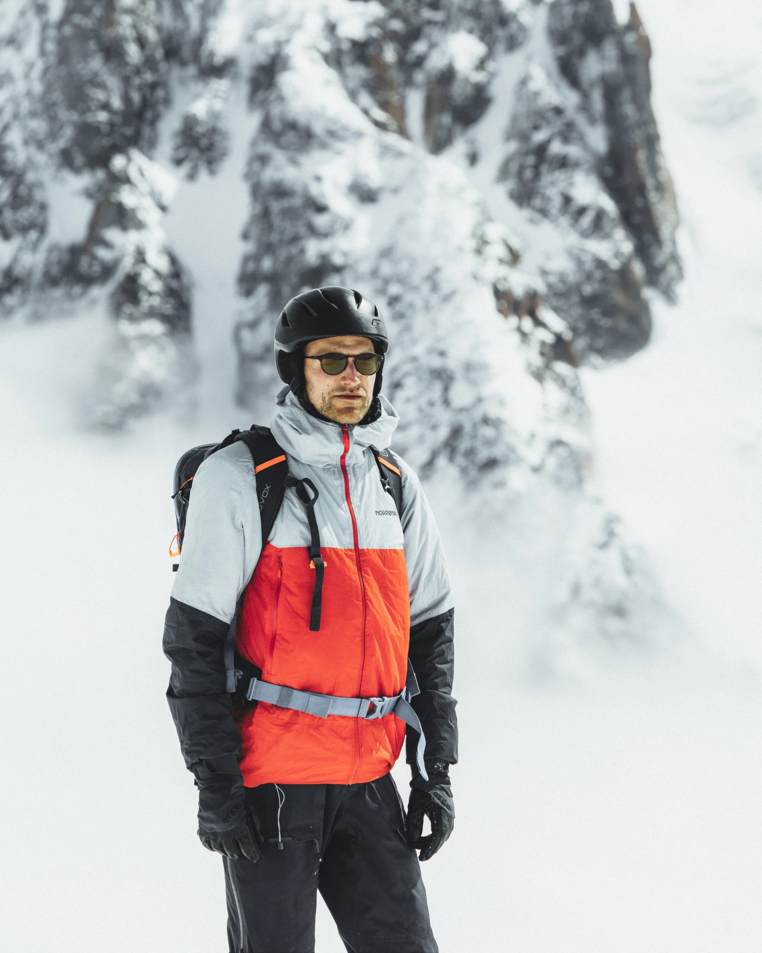 A man in winter gear, including a helmet, sunglasses, and a red and gray jacket, standing in a snowy mountain landscape.