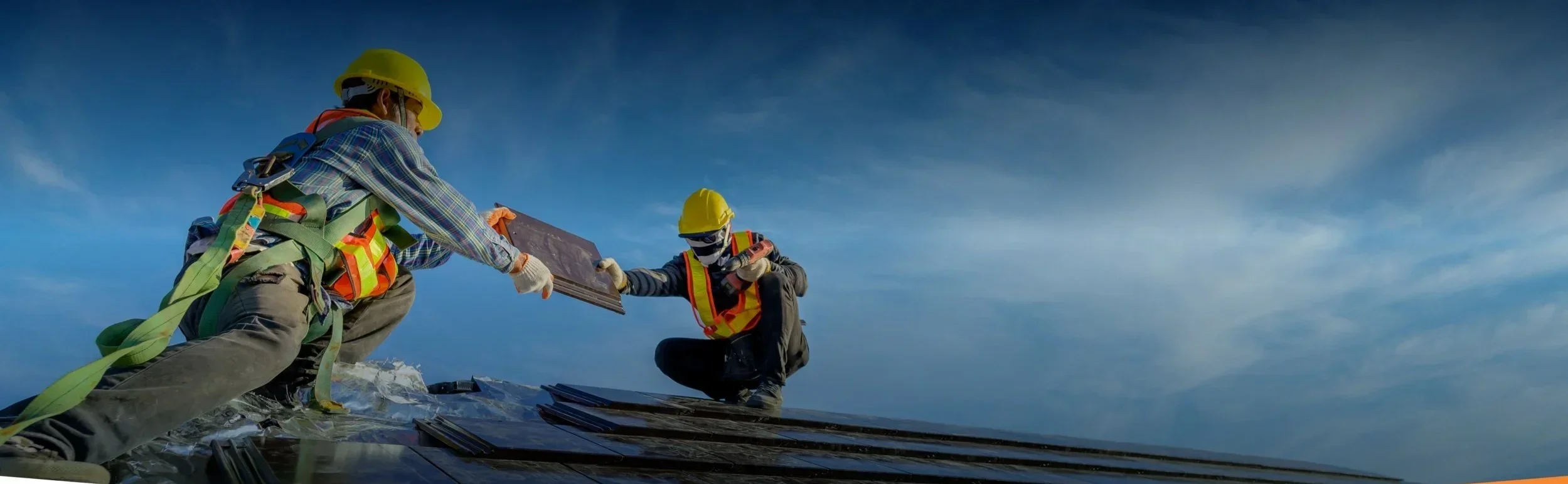 Two construction workers wearing yellow safety helmets and safety vests, installing solar panels on a roof during daytime with a cloudy sky.