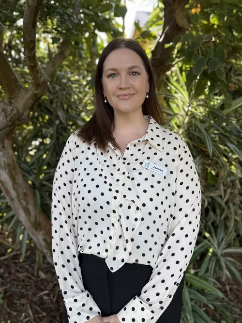 A young woman with long brown hair and pearl earrings standing outdoors in a garden with green foliage, wearing a white blouse with black polka dots and a name tag.