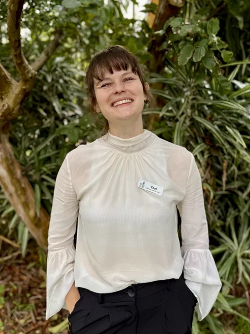 A woman with short brown hair and a big smile, wearing a white blouse and black pants, standing outdoors in front of green foliage.
