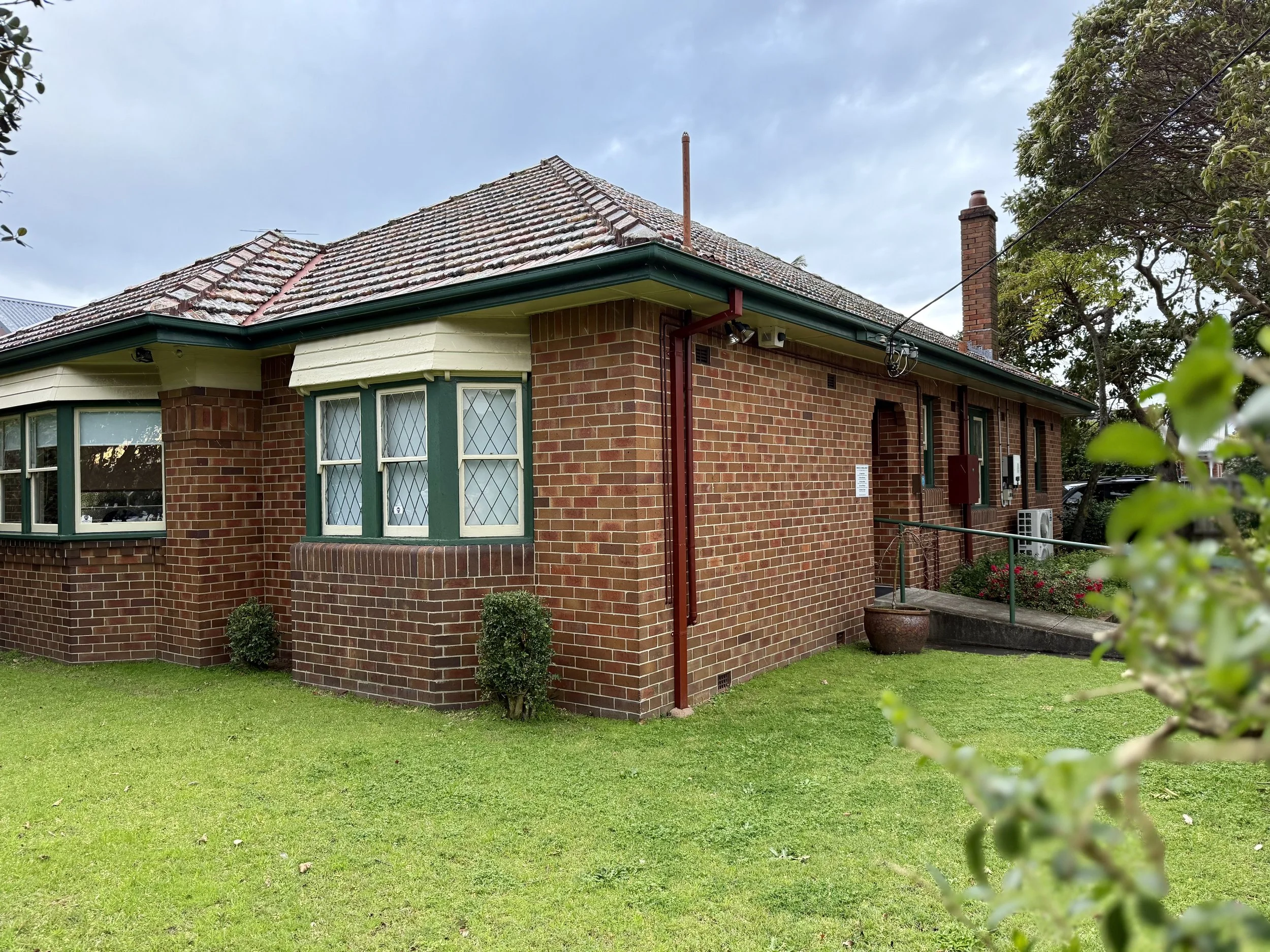 Red-brick house with green trim, bay window on the left, small shrubs, and a grassy yard under a cloudy sky.