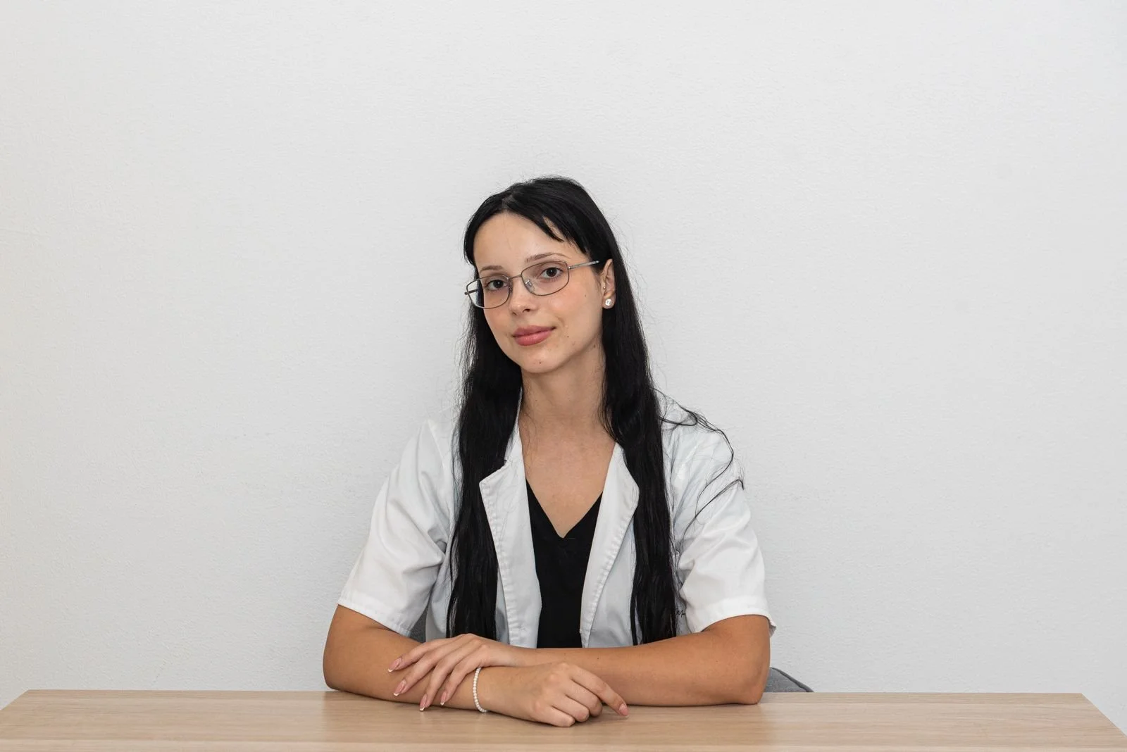 Mulher jovem com cabelos longos e pretos, usando óculos, roupa branca e blusa preta, sentada à mesa de madeira, com expressão neutra, contra parede branca.
