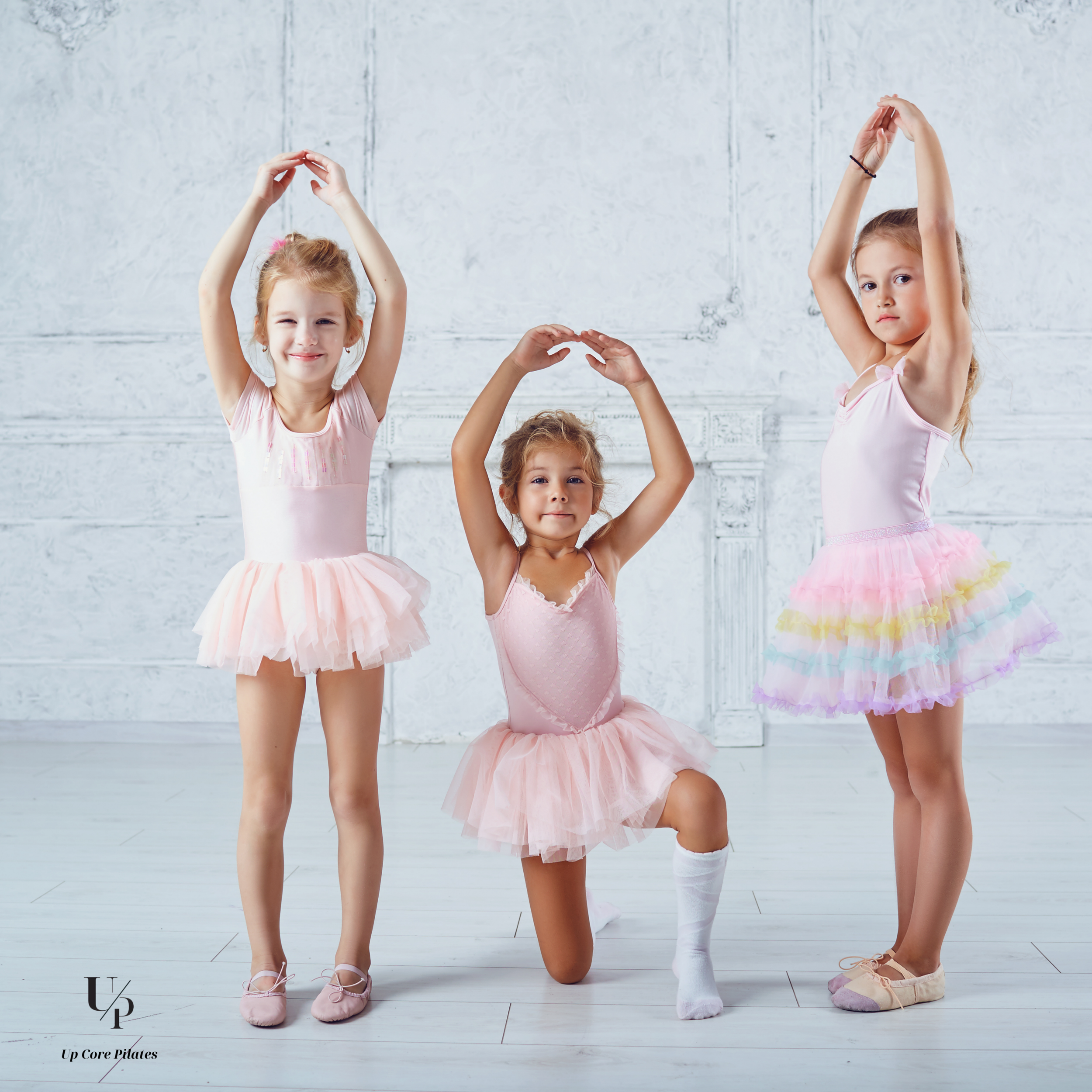 Three young girls dressed as ballerinas in pink tutus, practicing ballet moves in a dance studio with white walls.