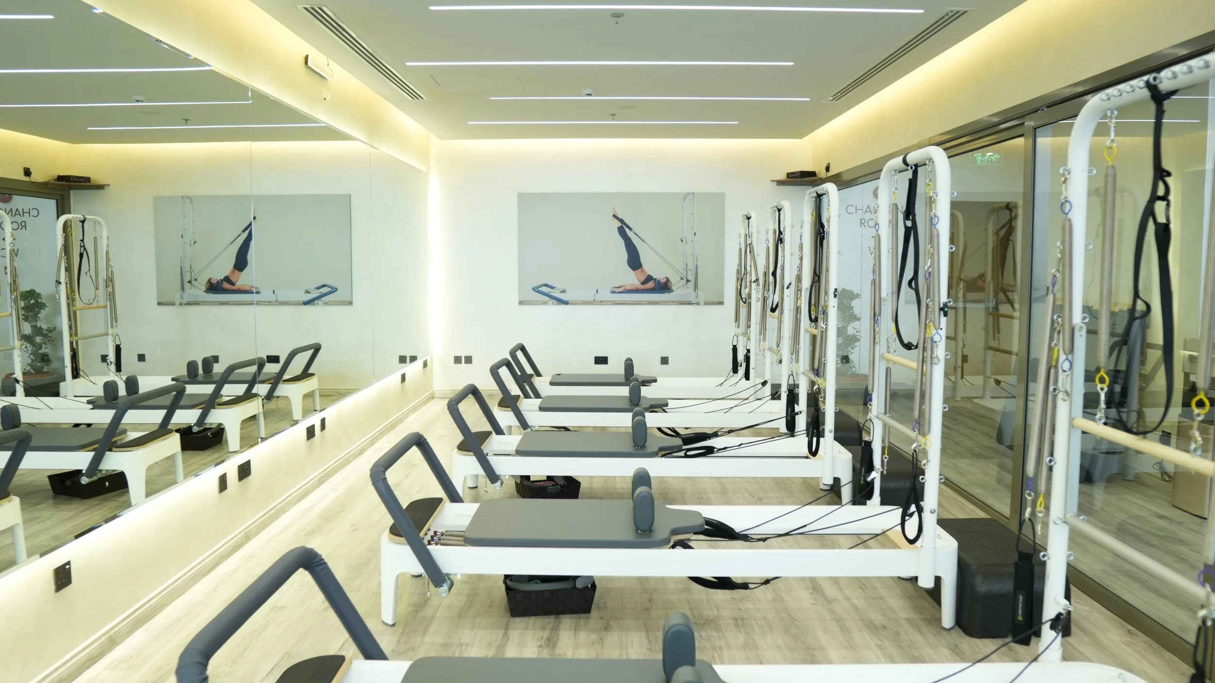 Pilates reformer machines in a brightly lit fitness studio with mirrored walls and a large wall mirror showing a woman exercising.